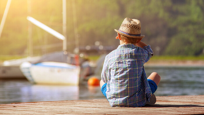© Soloviova Liudmyla, stock.adobe.com Ein kleiner Bub sitzt auf einem Holzsteg und blickt auf ein Segelboot