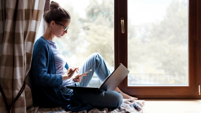 © Marina Andrejchenko, stock.adobe.com Junge Frau sitzt mit Smartphone und Laptop am Fenster